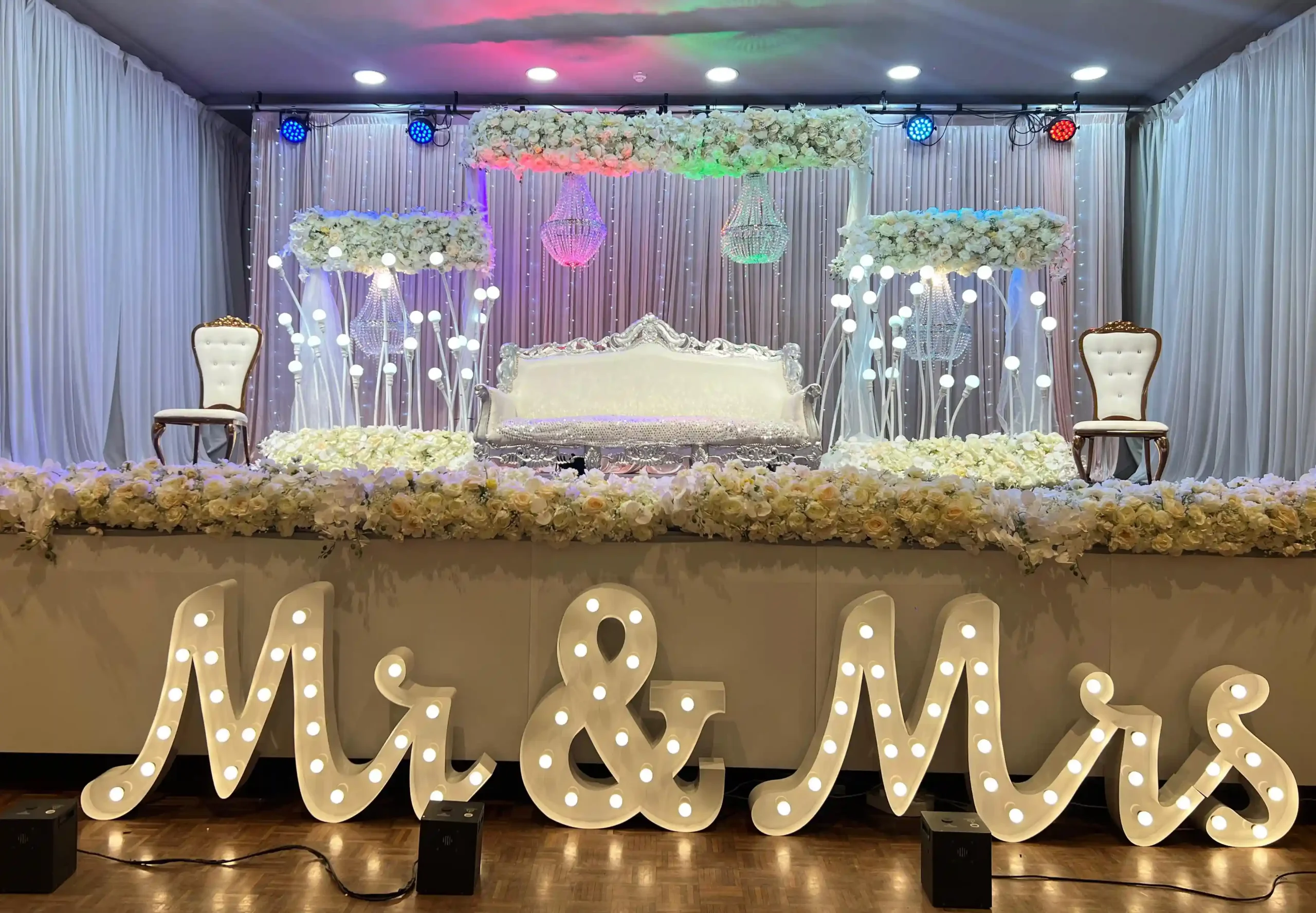 Decorated wedding stage with white sofa, flower arrangements, and illuminated Mr & Mrs letters.