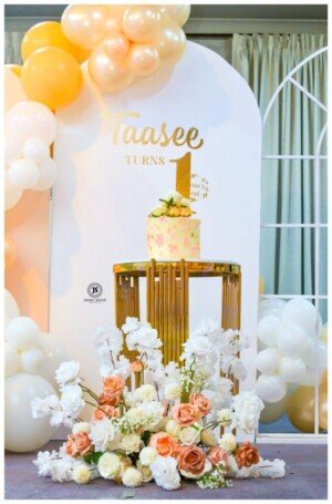 Birthday setup with cake on gold stand, peach balloons, and floral arrangements.