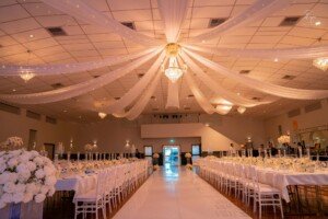 Banquet hall with long tables set, central white aisle, and ceiling fabric draping.