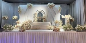 An event stage set with a white couch surrounded by white wall panels, a crystal chandelier arch, and blush pink and white floral arrangements.