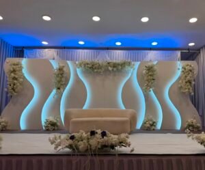 A modern event stage featuring wavy white backlit panels, white flower accents, and a beige sofa at the center.
