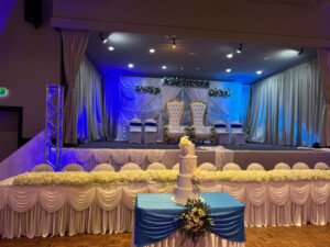 Event hall stage with blue lighting, white drapes, and a tiered cake.