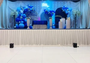 Decorated birthday party stage with blue balloons, backdrops, and number 16 display.