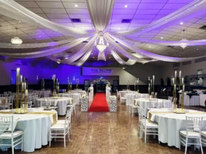 Decorated event hall with round tables, red aisle runner, purple lighting, and ceiling drapery.