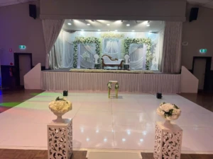 Decorated event stage with white couch, chairs, silver curtains, and white floral arrangements.