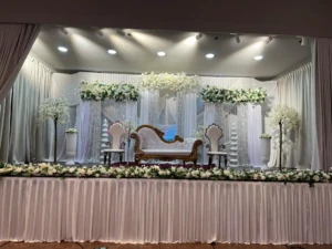 Decorated event stage with white couch, chairs, silver curtains, and white floral arrangements.