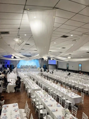 Banquet hall setup with long banquet tables, white chairs, and ceiling drapery.