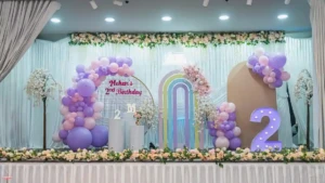Decorated 2nd birthday stage with pastel purple balloons, rainbow arches, and number 2 display.