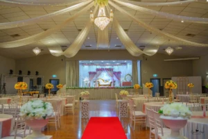 Decorated banquet hall with red aisle runner, yellow flower centerpieces, chandeliers, and stage.