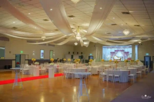 Decorated banquet hall with red aisle runner, yellow flower centerpieces, chandeliers, and stage.