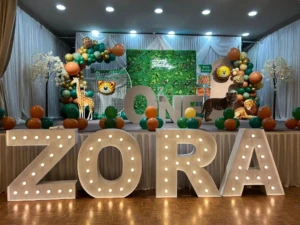 Jungle-themed birthday stage with lighted letters spelling ZORA, balloons, and animal decorations.