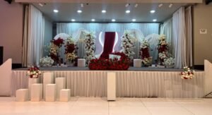 An event stage decorated with white curtain backdrops, white and red floral arrangements, and red rose decorations.