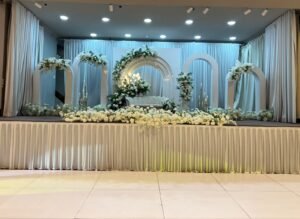 An decorated event stage featuring white archways, white flowers, draped curtains, and a white sofa at the center.