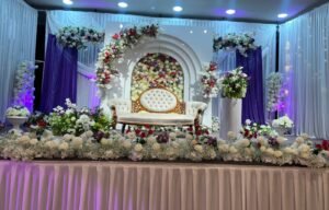 A white and gold loveseat set on a stage decorated with white and purple drapes, floral arches, and colorful flower arrangements.