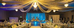 Banquet hall featuring ceiling drapes, chandelier lighting, and a blue-lit stage backdrop.